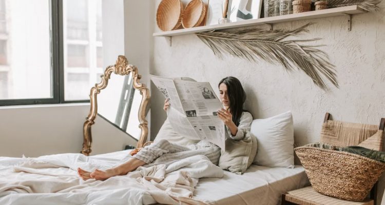 A woman comfortably reading a newspaper in a well-decorated, cozy bedroom setting.